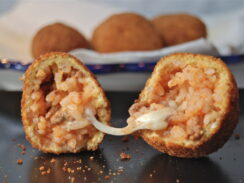A roman rice fritter, called suppli al telefono, sliced in half with a thread of mozzarella connecting the two halves. In the background, a white dish with more rice fritters.