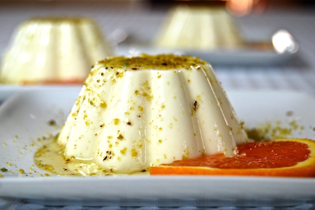 Panna cotta drizzed with ground pistachios and orange sauce on a square, white plate, with a orange slice as garnished. Two more panna cottas are in the background.