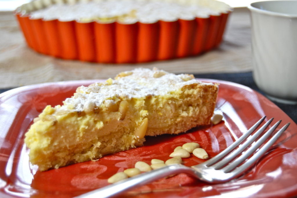 A slice of torta della nonna on a red plate with a fork and some pine nuts sprinkled on it. The entire torta della nonna in a red baking pan with fluted edges is in the background.