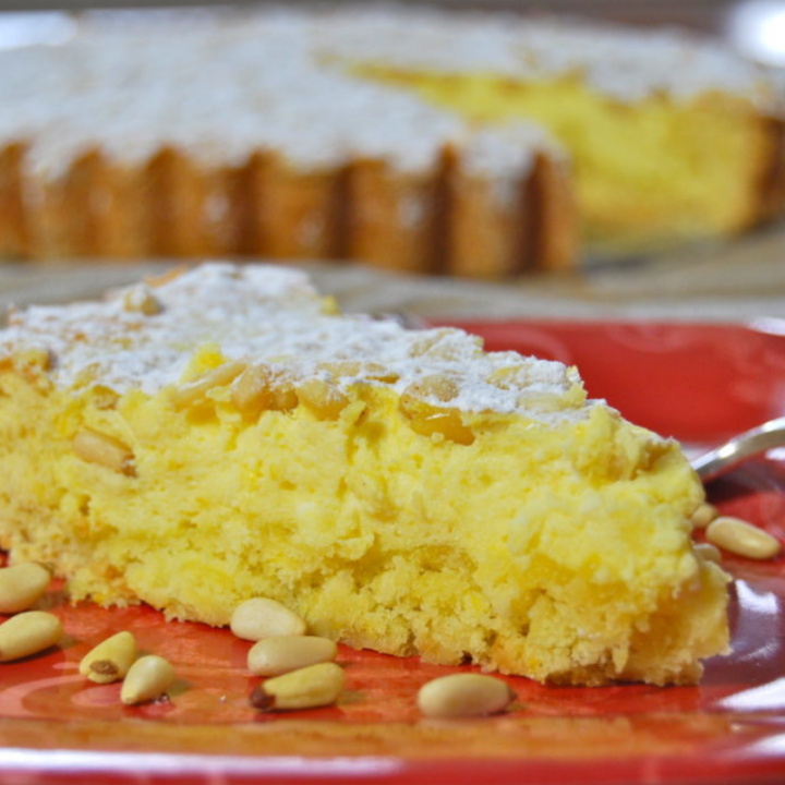 A slice of torta della nonna on a red plate with pine nuts sprinked on it. In the background is the entire torta della nonna with one slice missing.