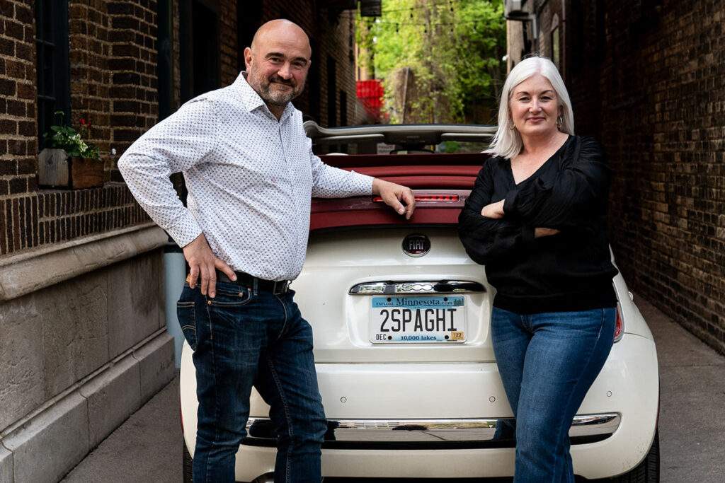Stefano and Cara of Due Spaghetti standing in an alley by their Fiat 500 with the license plate 2 SPAGHI