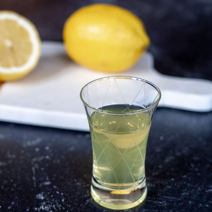 A glass of limoncello with lemons on a cutting board in the background.