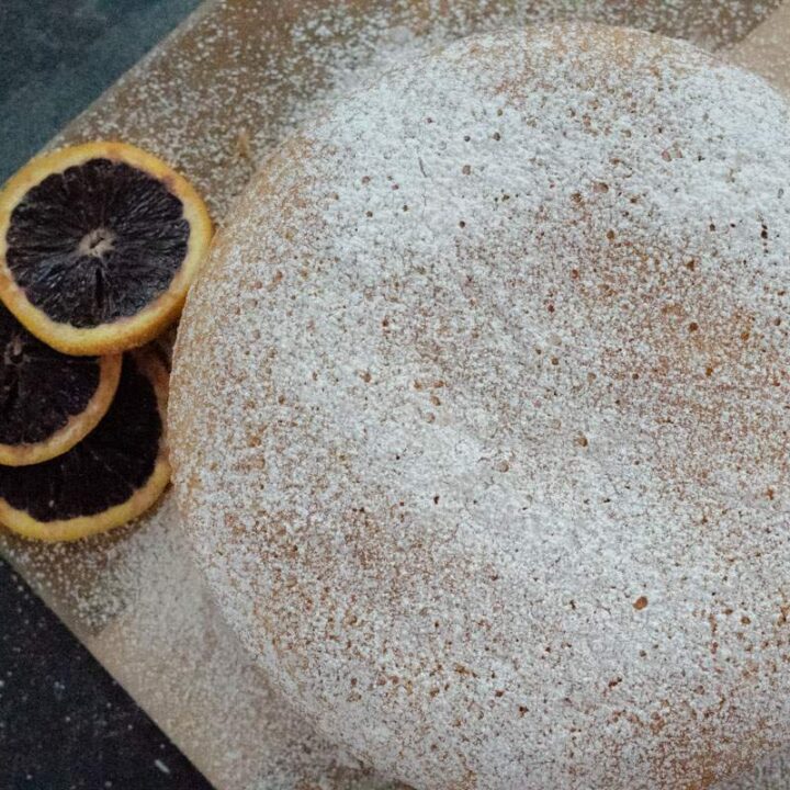 Olive oil cake on a cutting board with powdered sugar sprinkled on top and three slices of blood orange to the side.