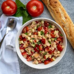 A bowl of panzanella with a spoon, onion, two tomatoes, bunch of basil and a baguette.