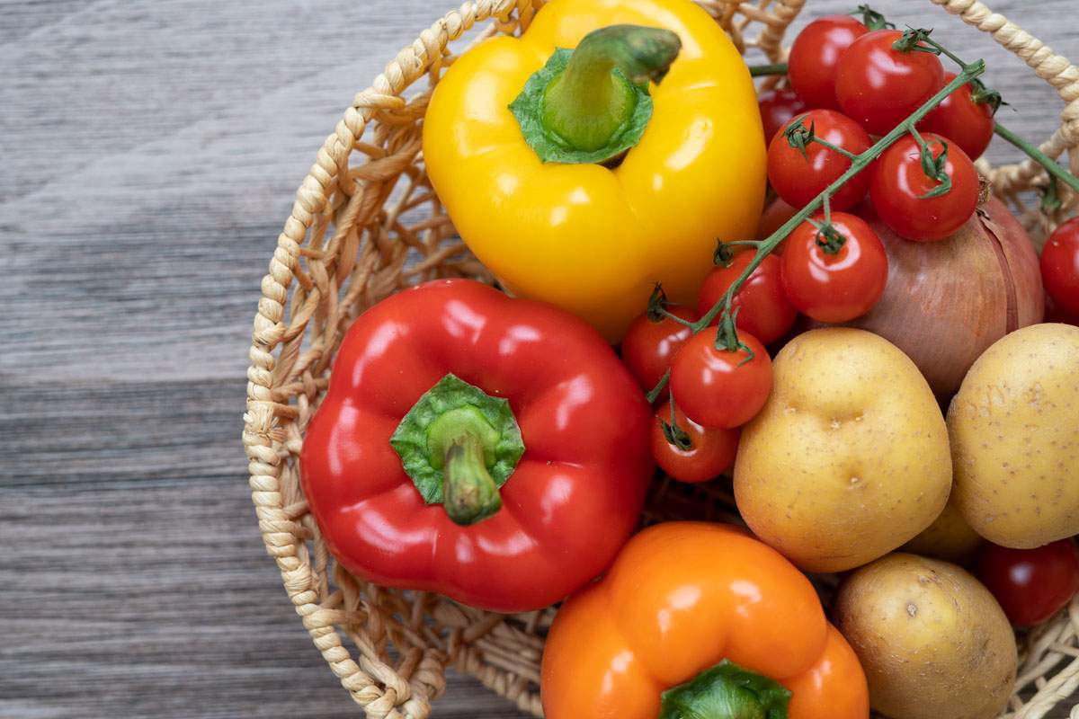 A basket with a red pepper, orange pepper, yellow pepper, potatoes, an onion and cherry tomatoes.