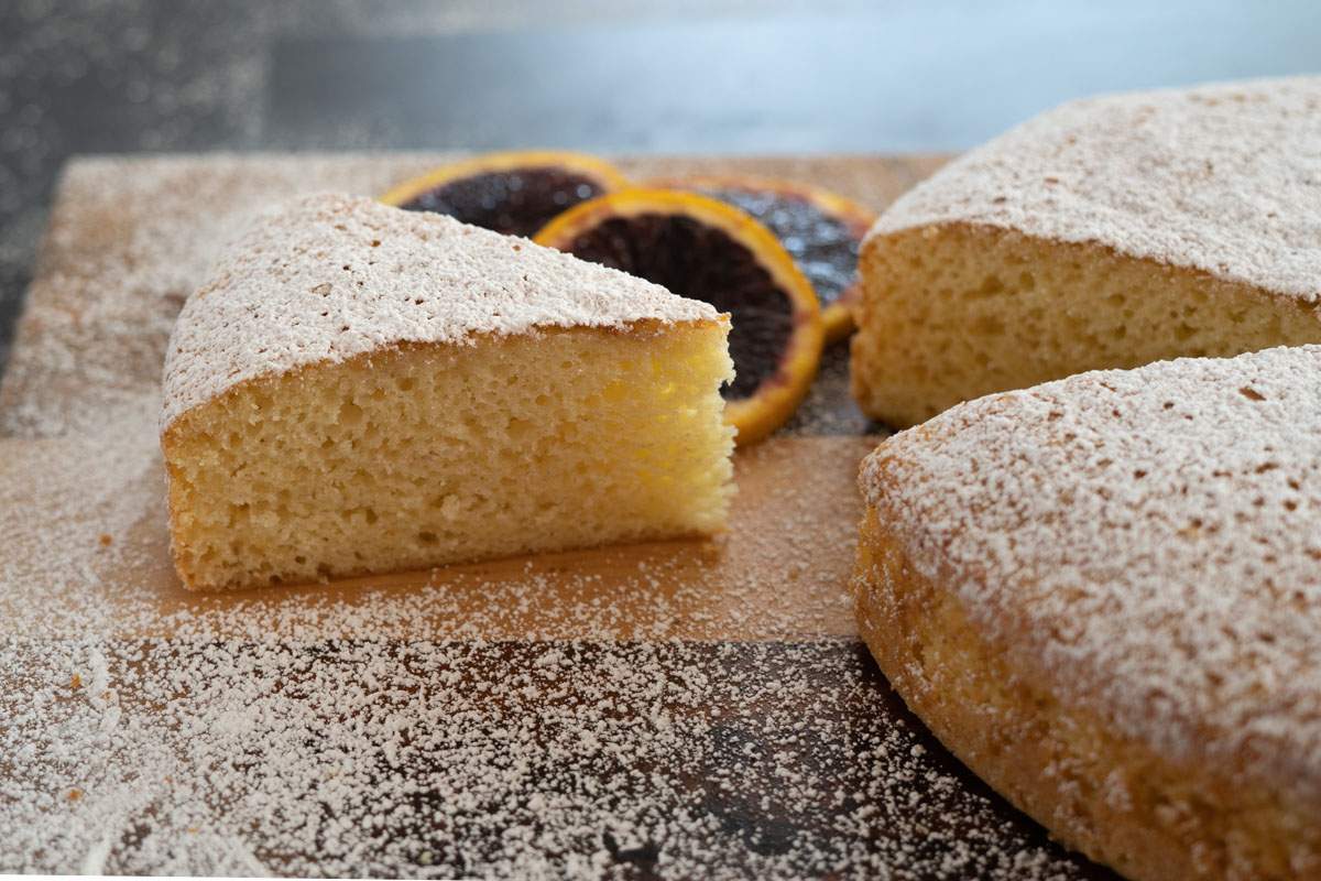 Olive oil cake with a slice pulled out on a cutting board sprinkled with powdered sugar and slices of blood oranges in the background.