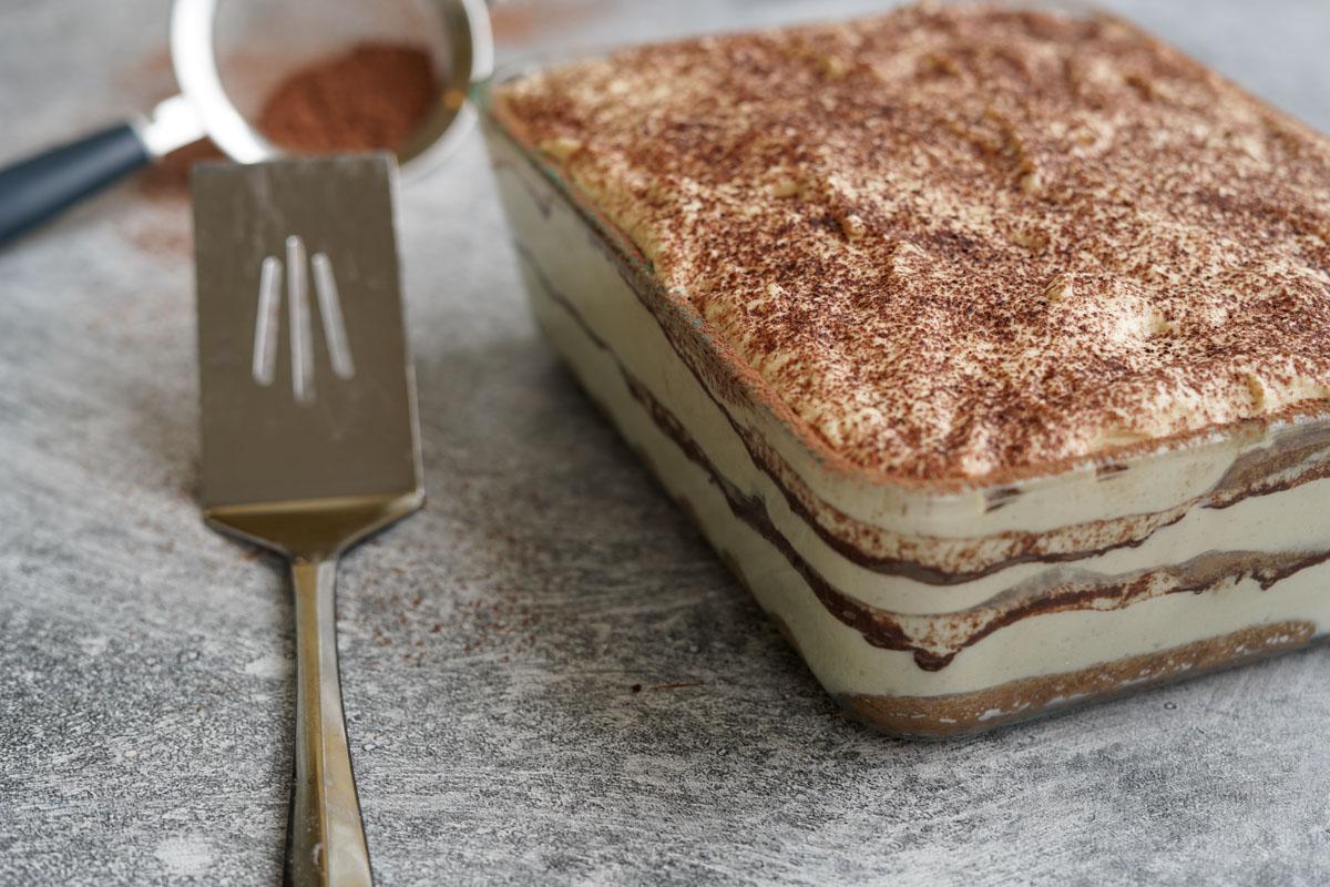 A tray of tiramisu with a stainless steel server next to it and a strainer with cocoa in the background
