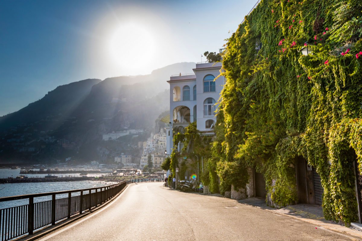 Amalfi drive leading to a village. A vine colored building is on the right and the sea is on the left.