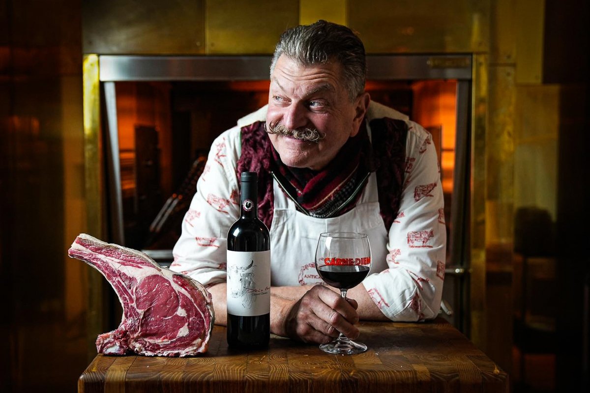 Butcher Dario Cecchini leaning on a table with a glass of red wine in his hand, and a bottle of wine and a piece of steak next to it.
