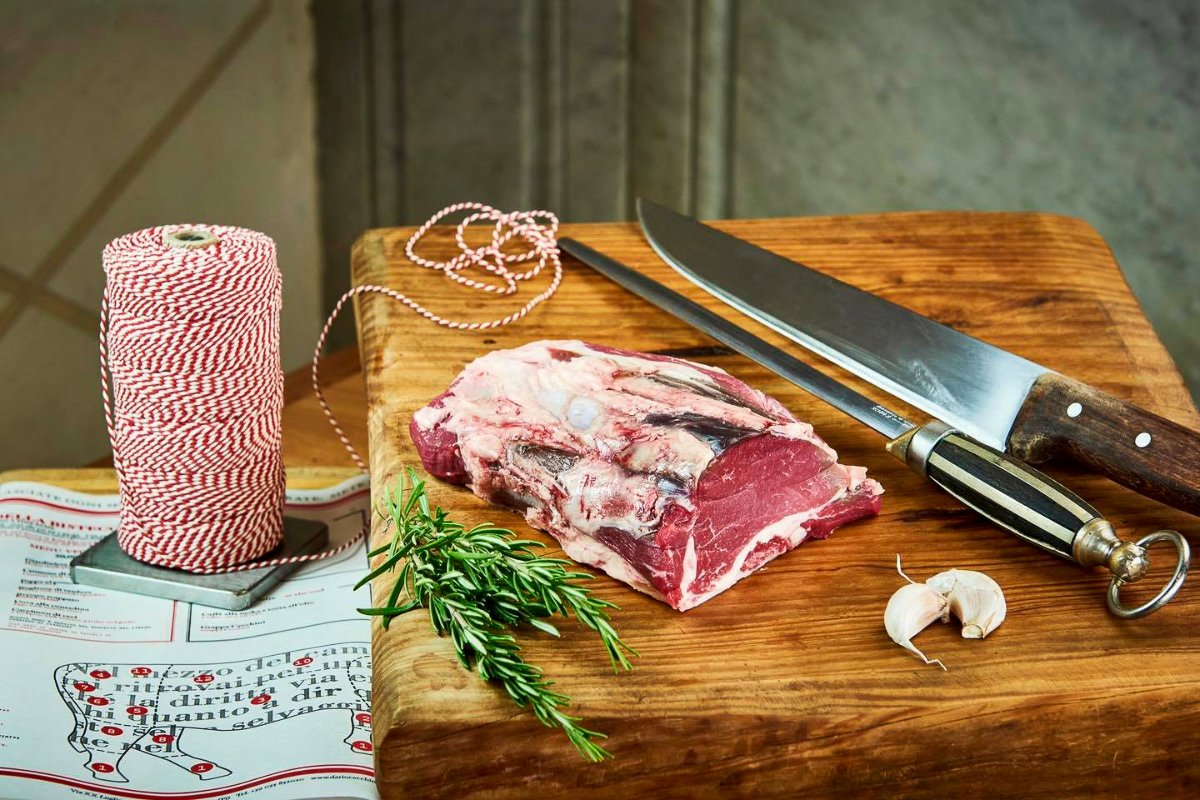 A piece of filetto on a piece of butcher block. To the right is a honing rod and a butcher knife. Two cloves of garlic are in the forefront and a spring of rosemary is to the left. On the table next to the butcher block is red adn white butcher twine.