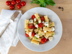 A plate of pasta fredda with a fork on a white napkin to the left, and some cherry tomatoes, a sprig of basil and some black peppercorns in the background.