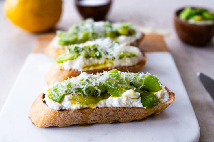 Three crostini with ricotta, fave and pecorino on a cutting board. In the background, there is a lemon, a small bowl of salt, another bowl of ricotta and fava beans, a few pieces of pecorino romano., a lemon, and a butter knife.