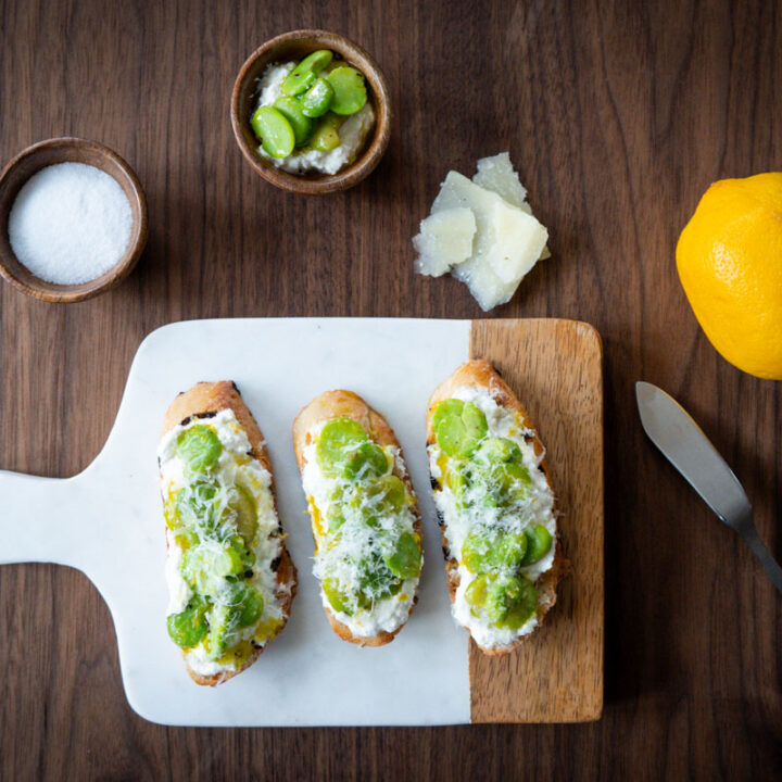 Three crostini with ricotta, fave and pecorino on a cutting board, surrounded by a small bowl of salt, another bowl of ricotta and fava beans, a few pieces of pecorino romano., a lemon, and a butter knife.